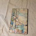 Handmade junk journal with coastal watercolor collage cover featuring seashells, treasure chest, driftwood, and dragonfly, closed with a navy elastic band