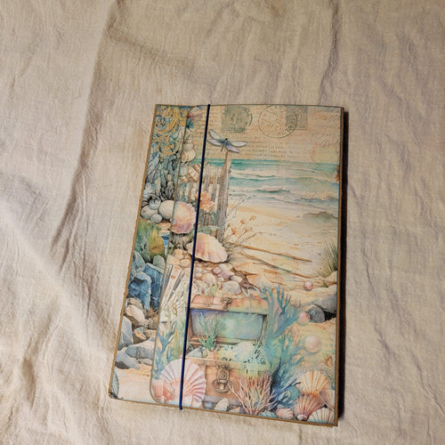 Handmade junk journal with coastal watercolor collage cover featuring seashells, treasure chest, driftwood, and dragonfly, closed with a navy elastic band